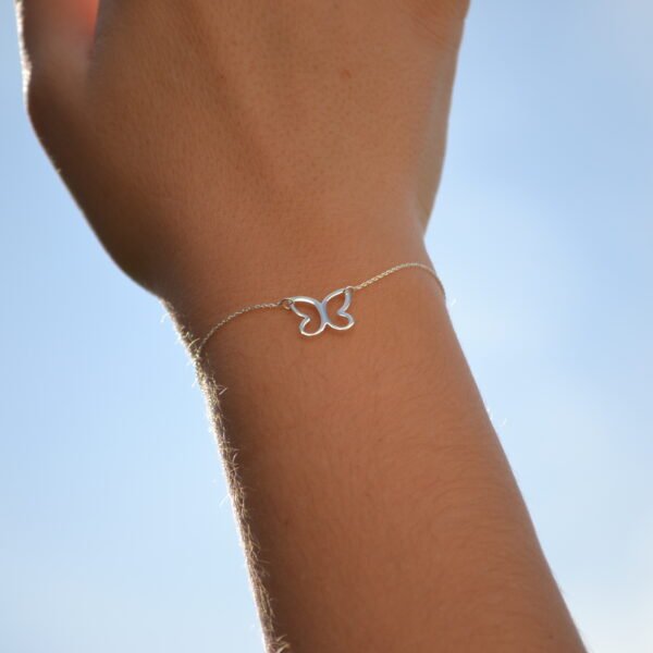 Mano alzada hacia el cielo luciendo una pulsera de plata de ley con forma de mariposa, símbolo de transformación y libertad. La mano tapa parcialmente el sol, creando un juego de luz natural que resalta el brillo de la pulsera y aporta una sensación de esperanza, renacer y conexión con la naturaleza. Imagen ideal para representar joyería artesanal de plata, pulseras con significado y accesorios emocionales inspirados en el cambio y la luz.